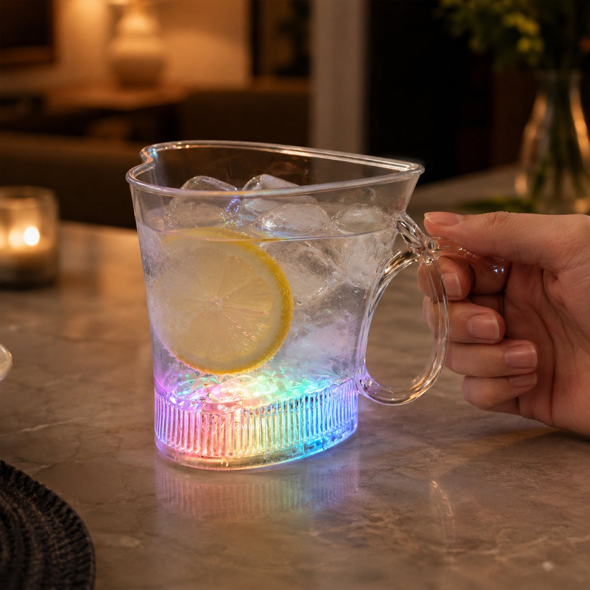 A hand holds a clear heart-shaped mug with a lemon slice in water, its base glowing with rainbow colors.