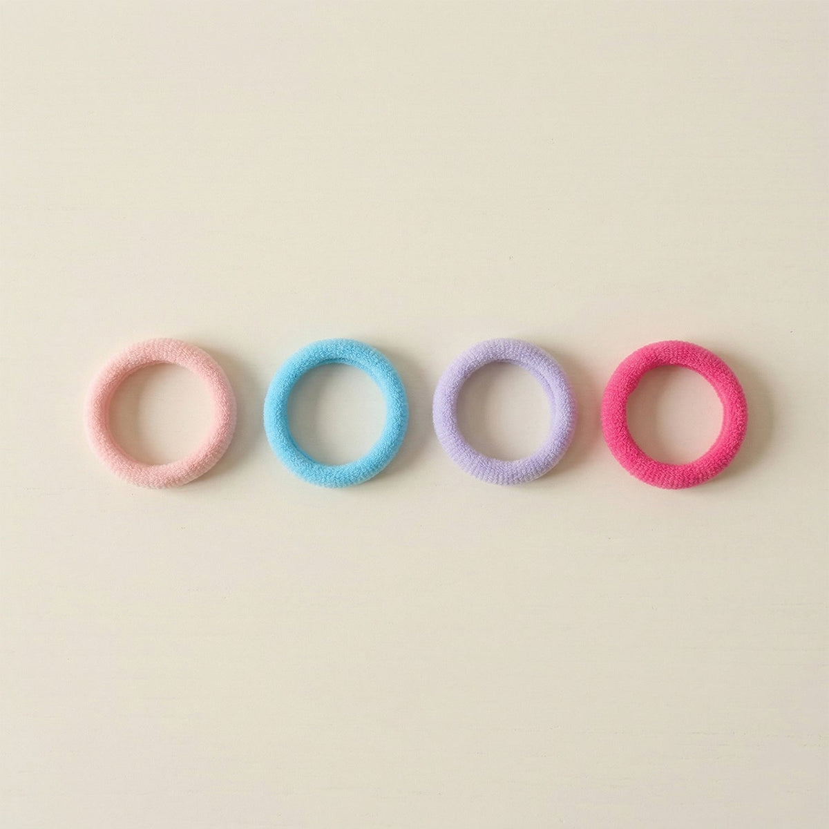 Four colorful elastic hair ties in pink, blue, lavender, and fuchsia arranged in a row on a light beige surface.