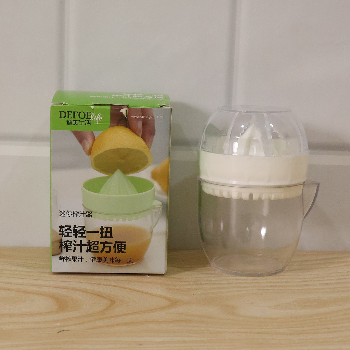A boxed citrus juicer next to the actual white and clear plastic manual citrus squeezer on a wooden surface.