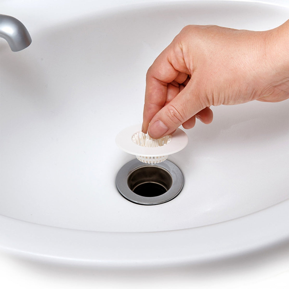 A hand holds a white plastic sink drain strainer above a sink's metal drain opening.