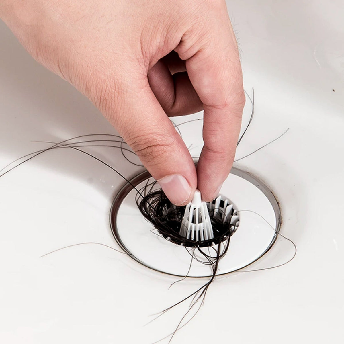 A hand removes a white plastic sink drain strainer from a drain, pulling out a clump of dark hair.