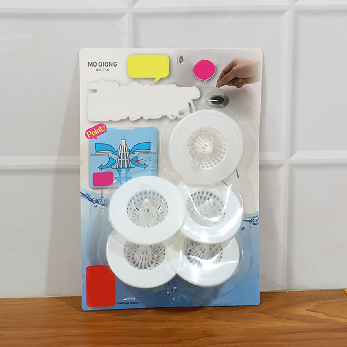 A blister pack containing five white sink drain strainers is displayed on a wooden surface.