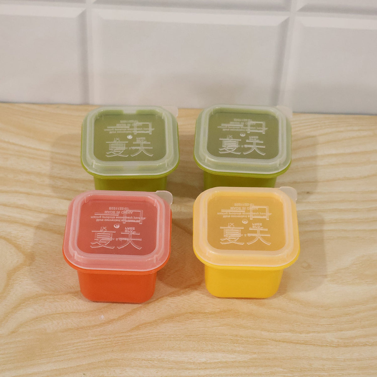 Four square silicone molds in green, red, and yellow with clear lids, arranged on a light wooden surface against a white