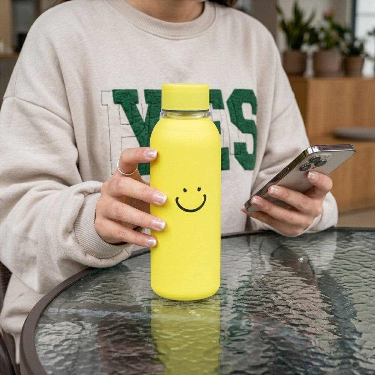 A person holding a yellow glass water bottle with a smiley face and a smartphone at an outdoor table.