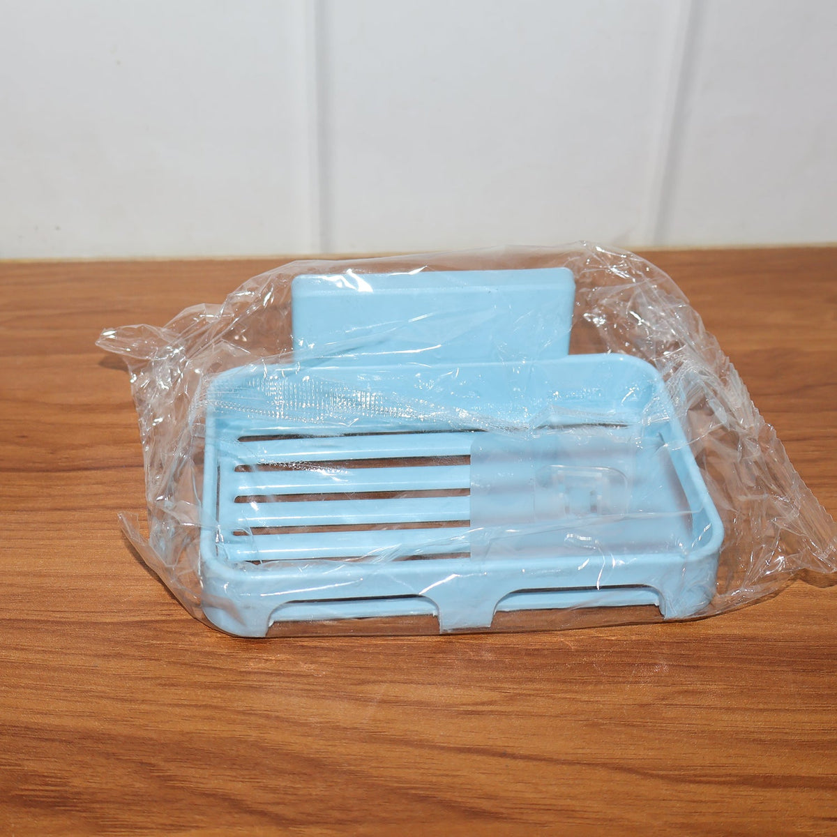 Light blue plastic soap dish wrapped in clear plastic packaging, resting on a wooden surface.