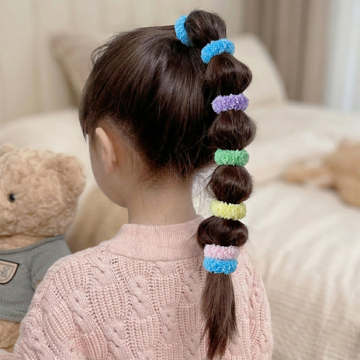 A child's long dark hair is styled with multiple colorful fluffy elastic hair bands creating a segmented ponytail.