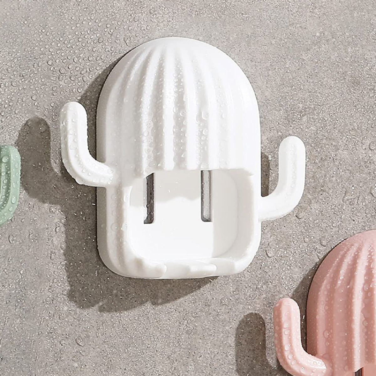 White cactus toothbrush holder covered in water droplets, mounted on a wet gray tiled wall next to a pink one.