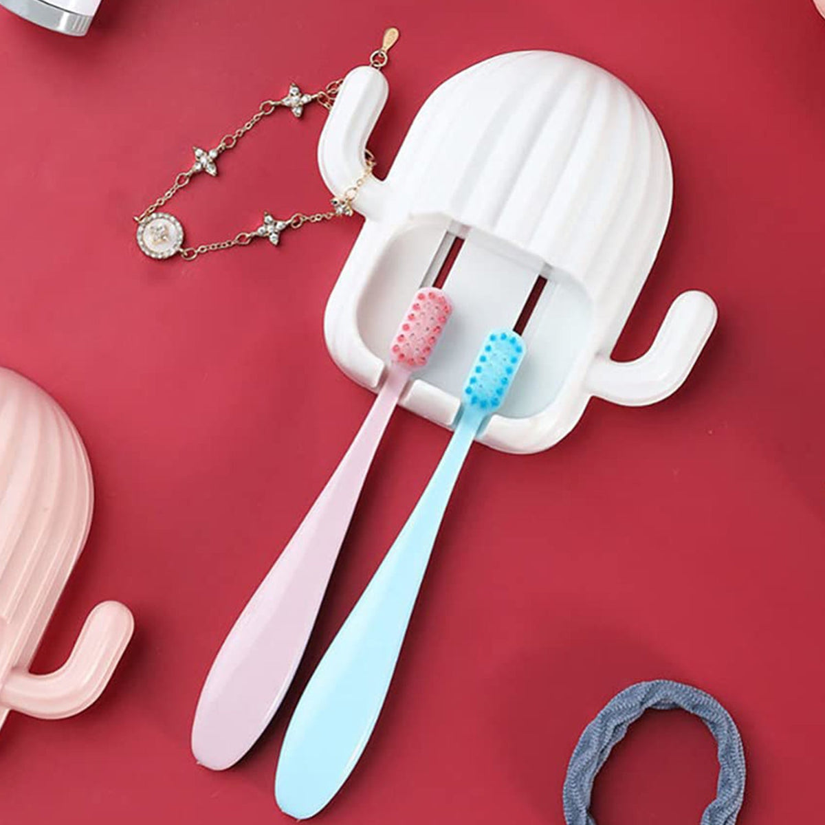 White cactus toothbrush holder on a red surface, holding pink and blue toothbrushes, with a bracelet nearby.