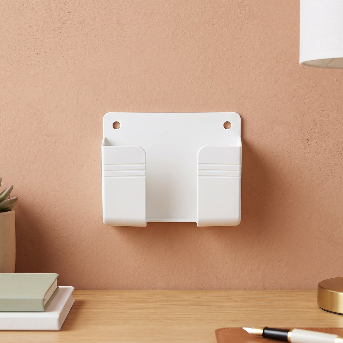A white wall-mounted phone holder with two compartments on a peach-colored wall above a wooden desk.