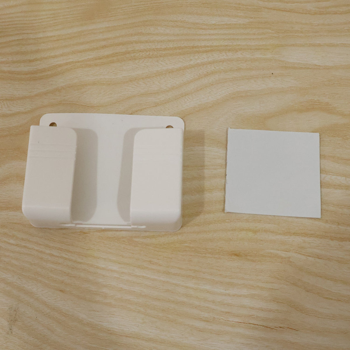 A white wall-mounted phone holder and a small white square adhesive pad on a light wooden surface.