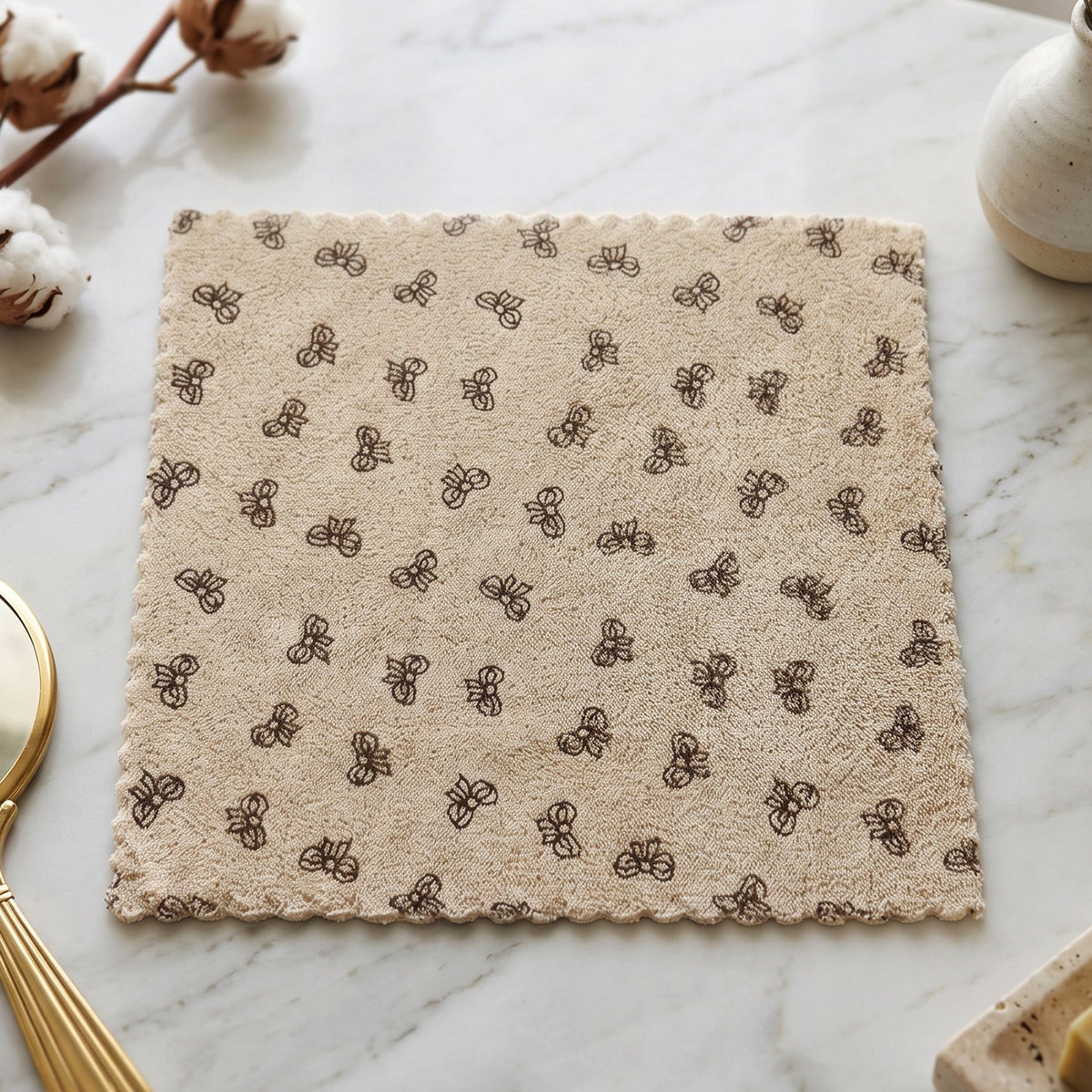 A beige microfiber towel with a dark bow pattern and scalloped edges lies flat on a white marble surface, with cotton