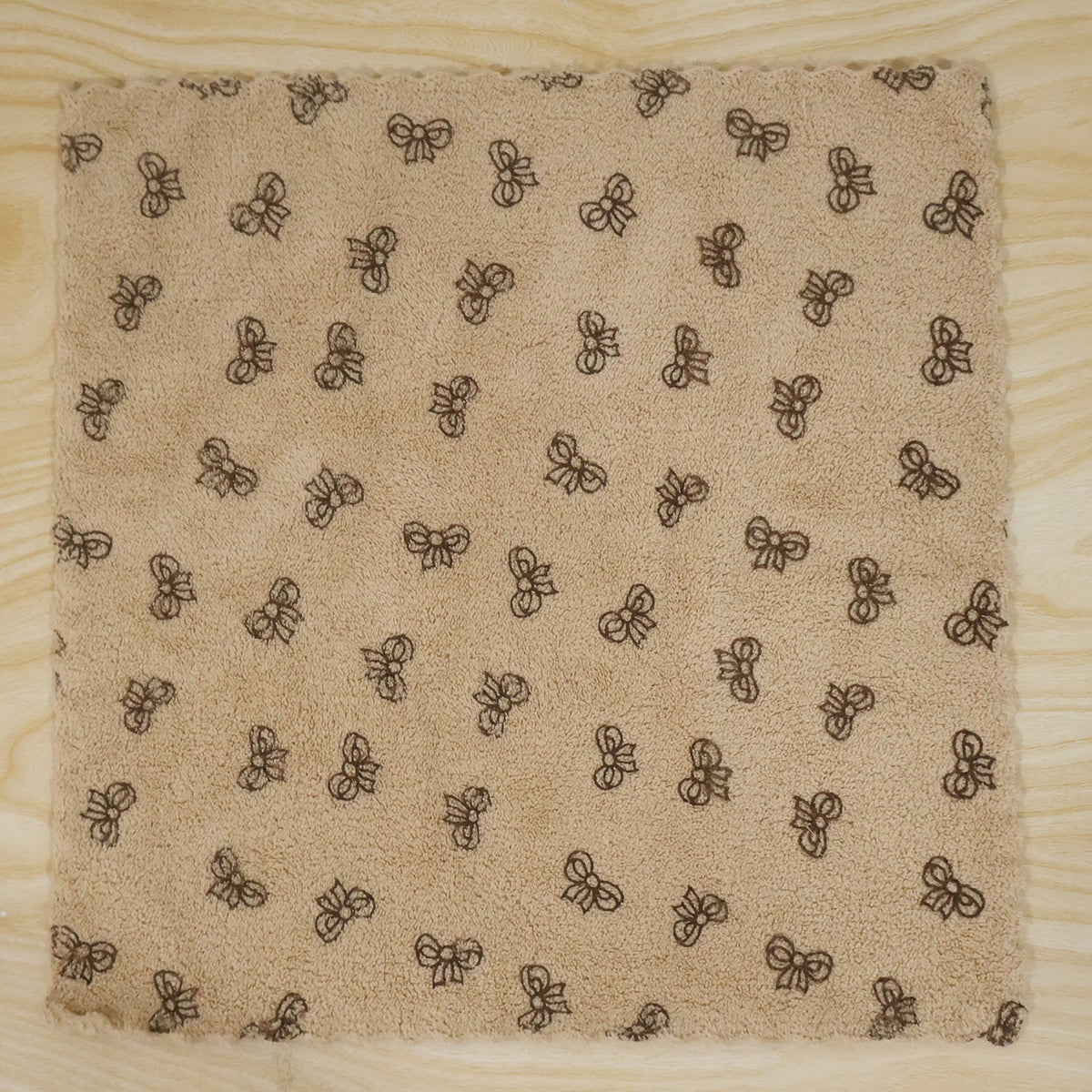 A beige microfiber towel with a dark bow pattern and scalloped edges lies flat on a light wood surface.
