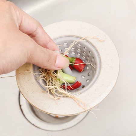 A hand placing vegetable scraps, including roots and chili, into a beige and grey silicone sink strainer.